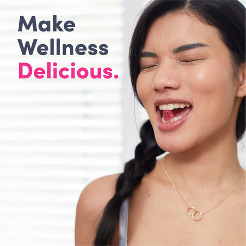 Young adult woman with dark hair and closed eyes, mouth open with a Biotin gummy inside. Graphic text: 'Make Wellness Delicious.'